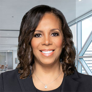 The image shows Traci Bransford in business attire, smiling confidently. She is standing in a modern office setting with large windows and a sleek, contemporary design. The lighting highlights her features, emphasizing professionalism and approachability.