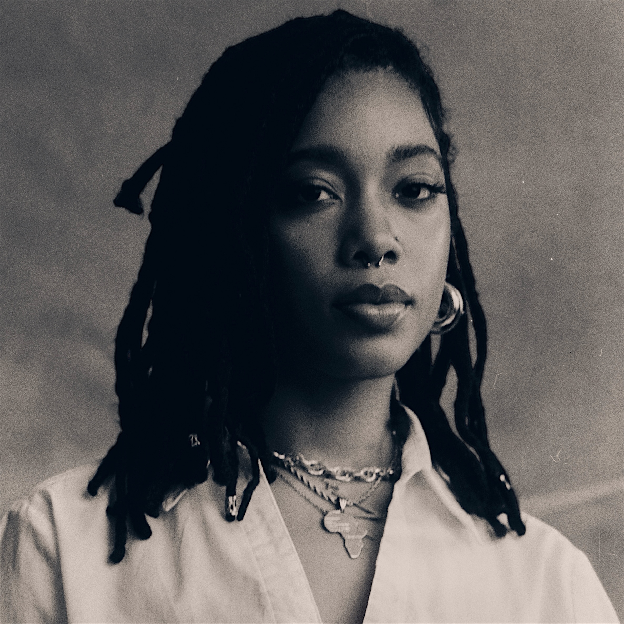 A grayscale portrait of Naima Eggleston with locs, wearing a nose ring and multiple necklaces, including one with a pendant shaped like Africa. She has a serene and confident expression, with hoop earrings visible. The photo has a soft, vintage aesthetic.